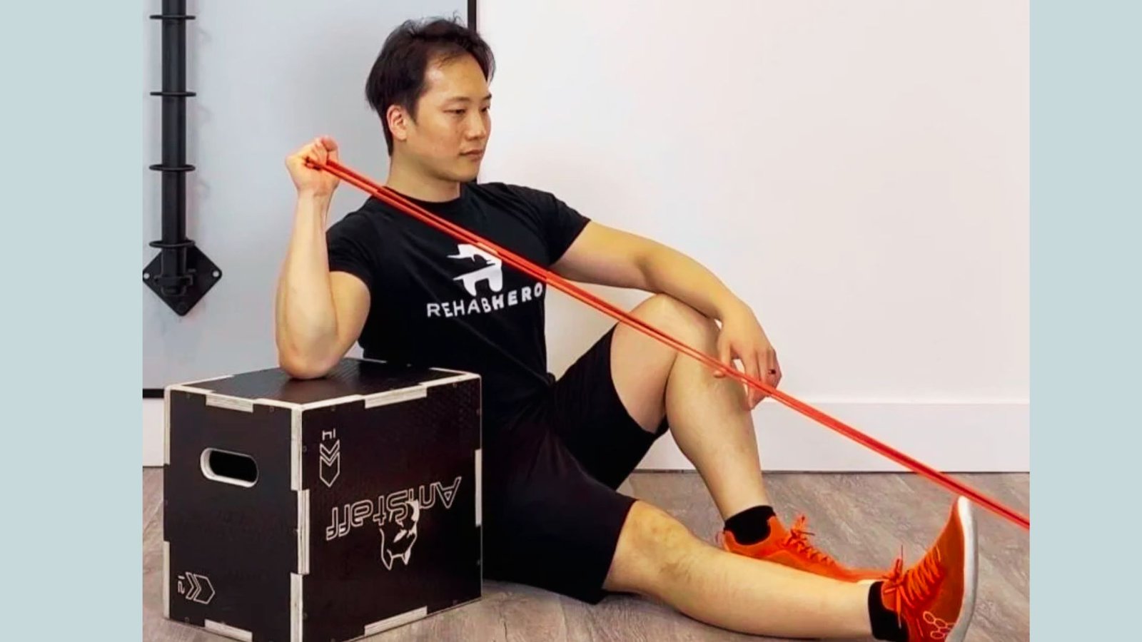 Man in black RehabHero shirt doing seated external rotation with red resistance band on plyo box.