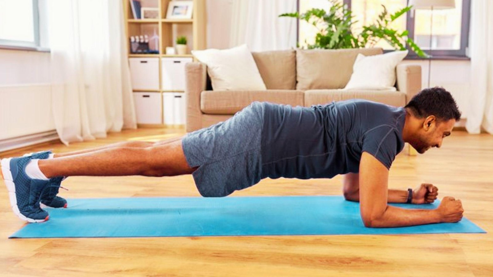 Man in dark gray shirt holding forearm plank on blue yoga mat in bright living room with couch.