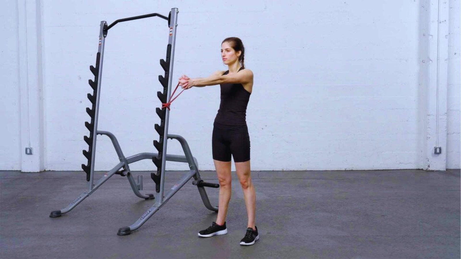 Woman doing standing resistance band chest press anchored to squat rack for upper body strength.