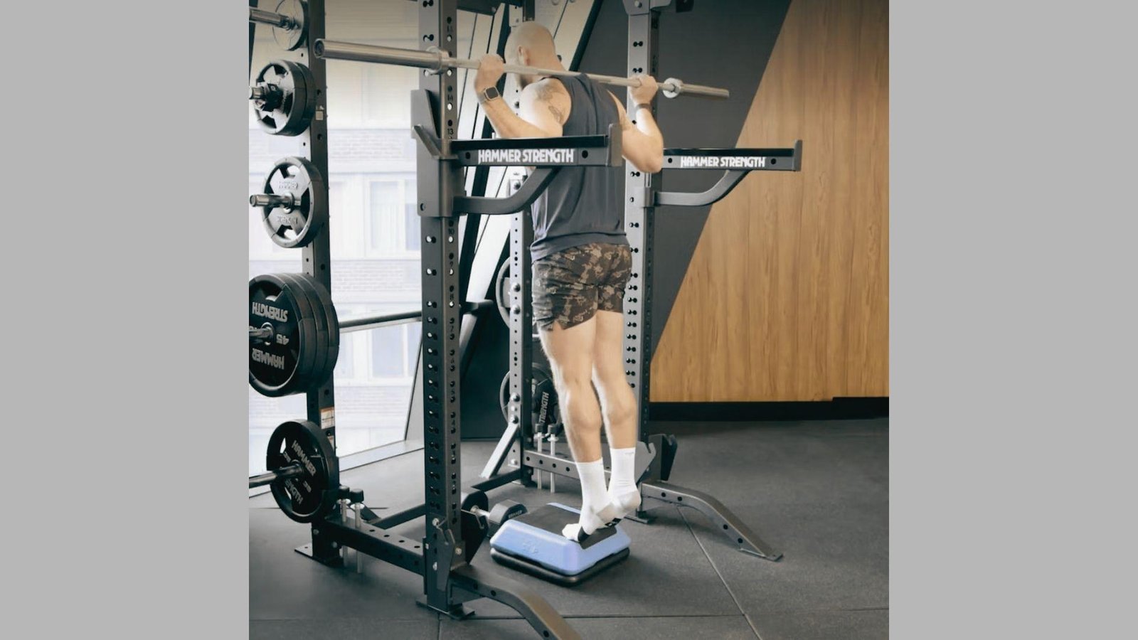 Man in dark tank and camo shorts doing standing barbell calf raises on blue step inside squat rack.