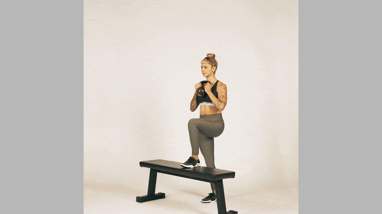Tattooed woman in black sports bra and gray leggings doing kettlebell step-up on black bench.