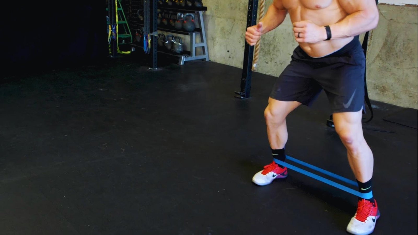 Shirtless man doing lateral band walk with blue long resistance band around ankles for glutes.