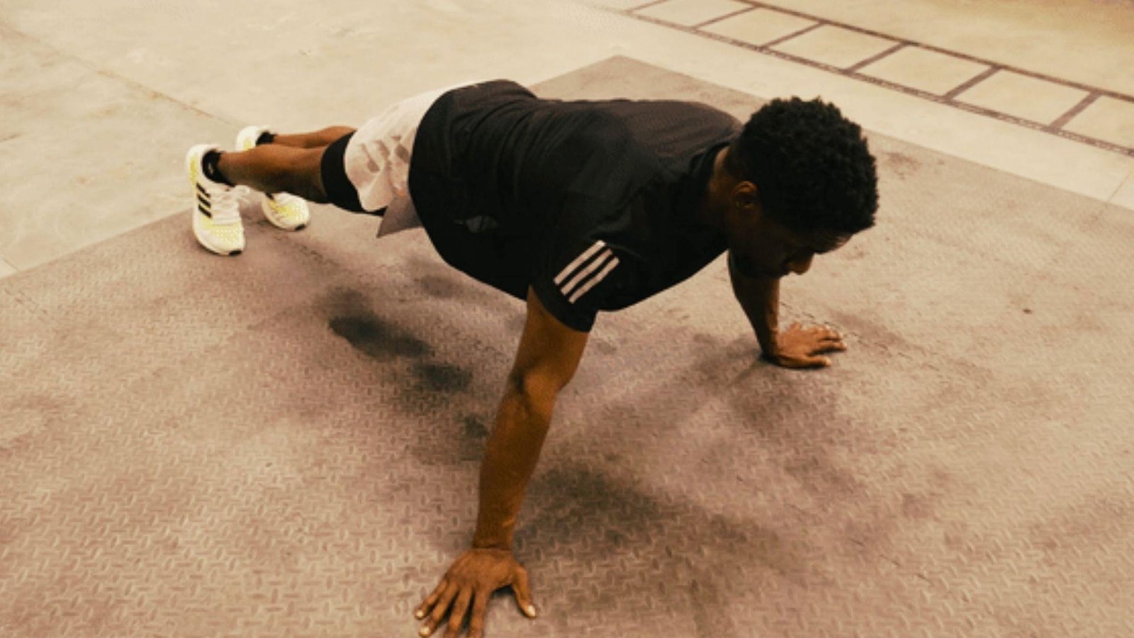 Man in black shirt doing wide-arm push-up plank on textured gym floor wearing white and yellow sneakers.