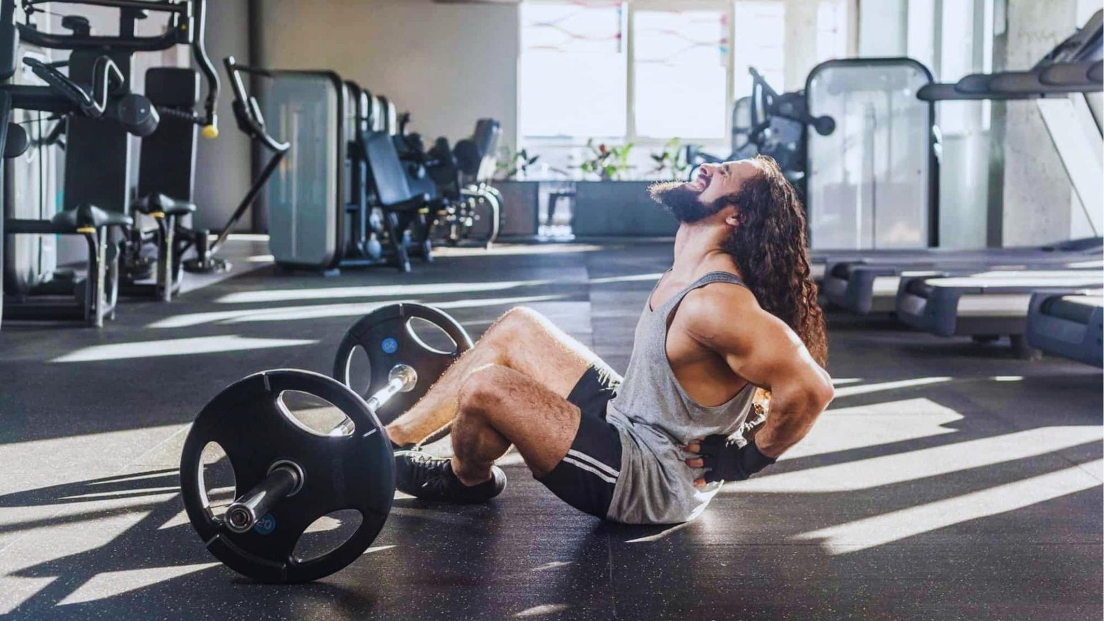 Man suffering lower back injury sitting on gym floor clutching back next to barbell after squat.