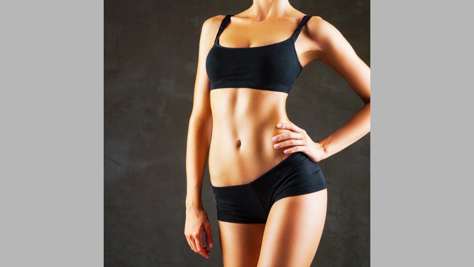 Fit woman from neck down in black sports bra and shorts with hand on hip against dark background.