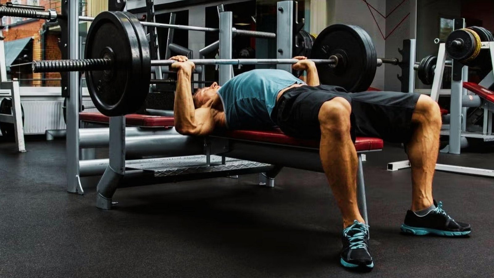 Muscular man in blue shirt bench pressing heavy barbell with large black plates in gym.