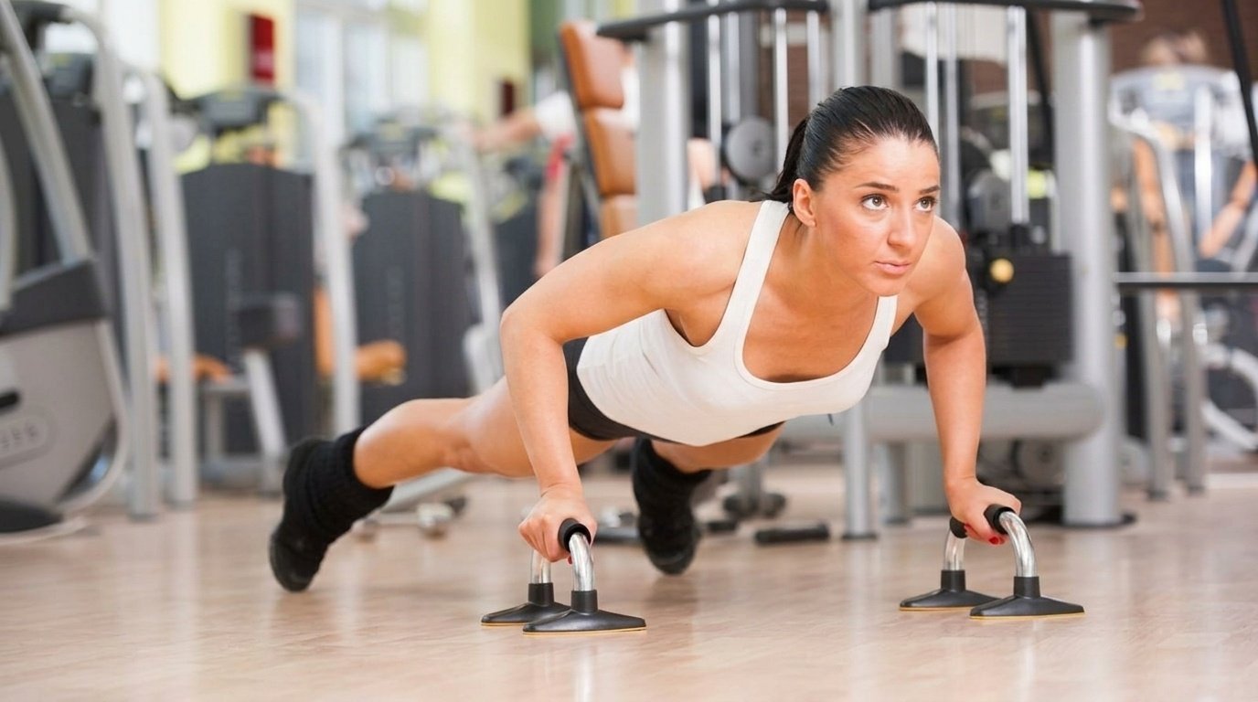 Woman in white tank top doing push ups on handle bars in a gym.