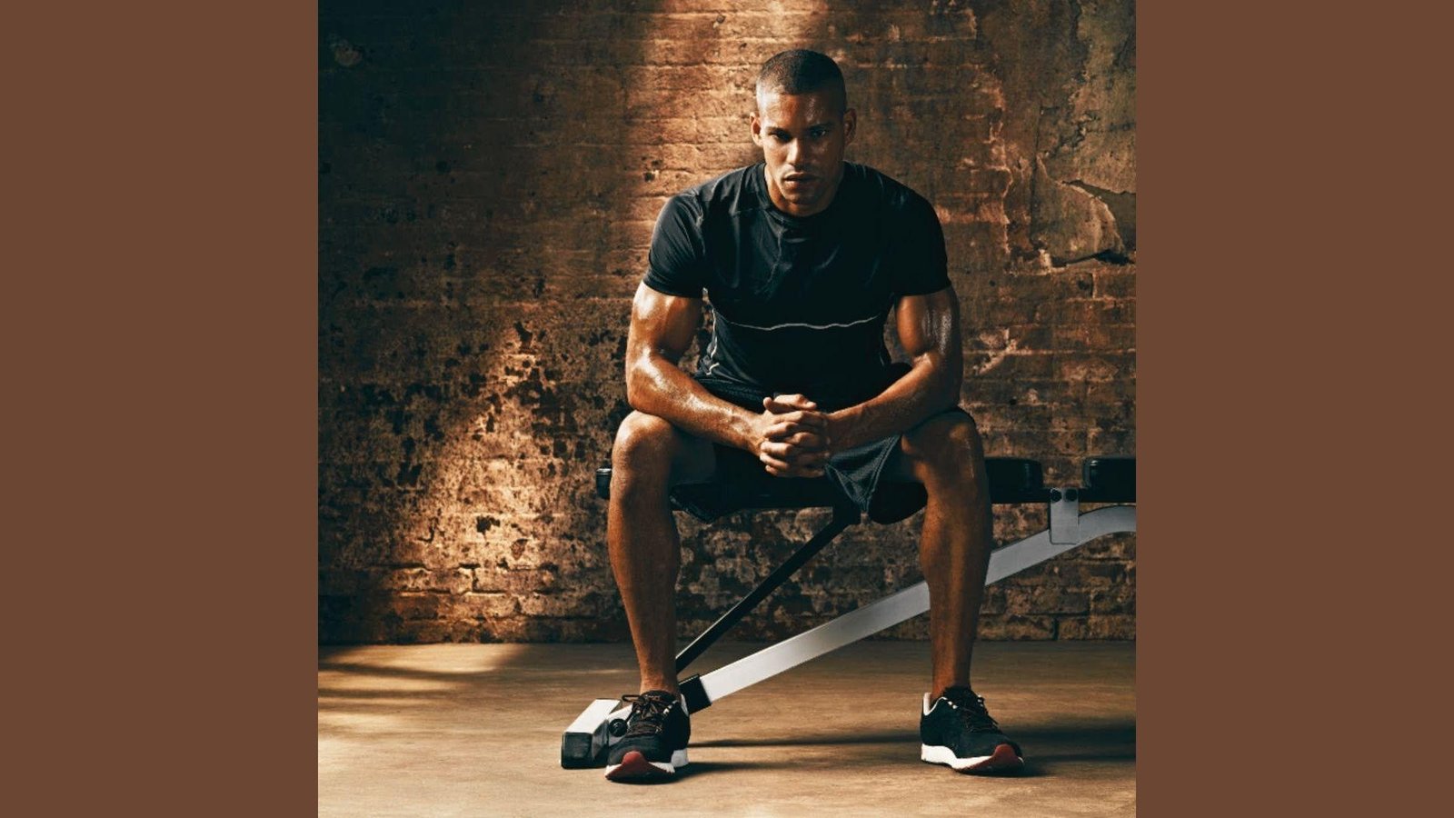 Muscular man in black shirt and shorts sitting on gym bench leaning forward, sweaty, brick wall behind.