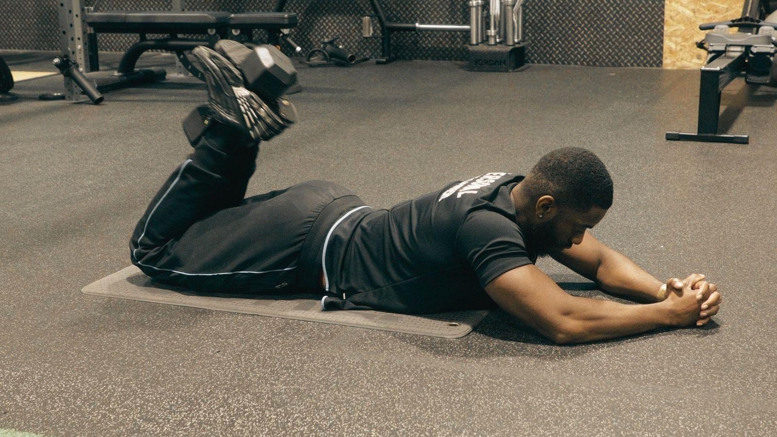 Man in black shirt and pants lying prone on floor mat doing dumbbell hamstring curl holding weight with feet.