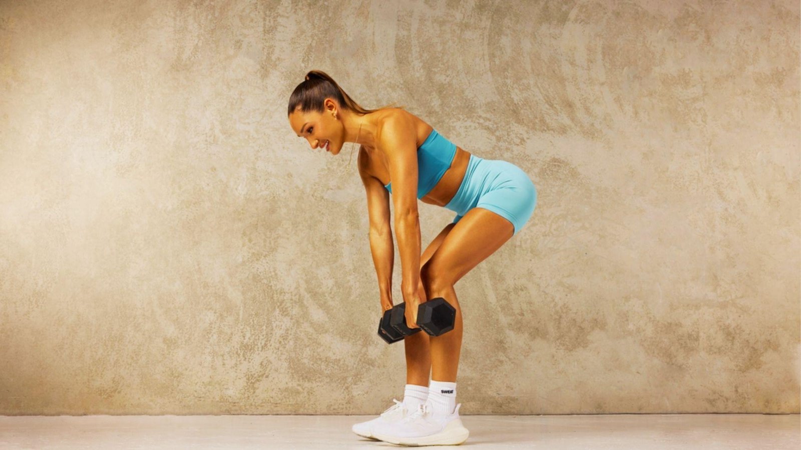 Fit woman in light blue sports bra and shorts doing dumbbell Romanian deadlift against textured beige wall.