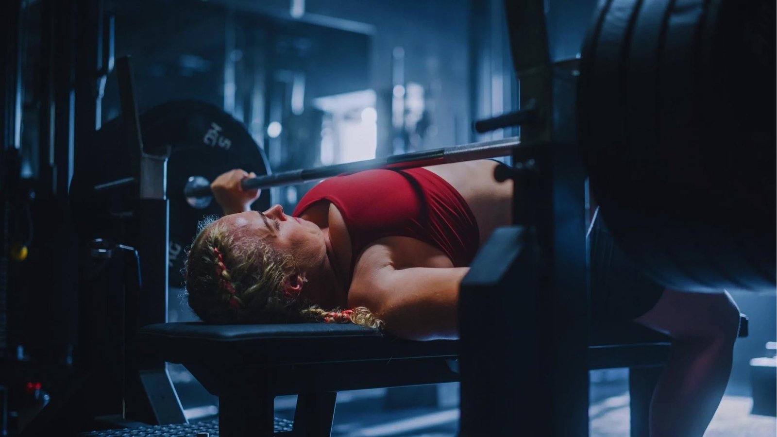 Woman in red sports bra doing heavy barbell bench press in dark gym, shoulder pain when benching.