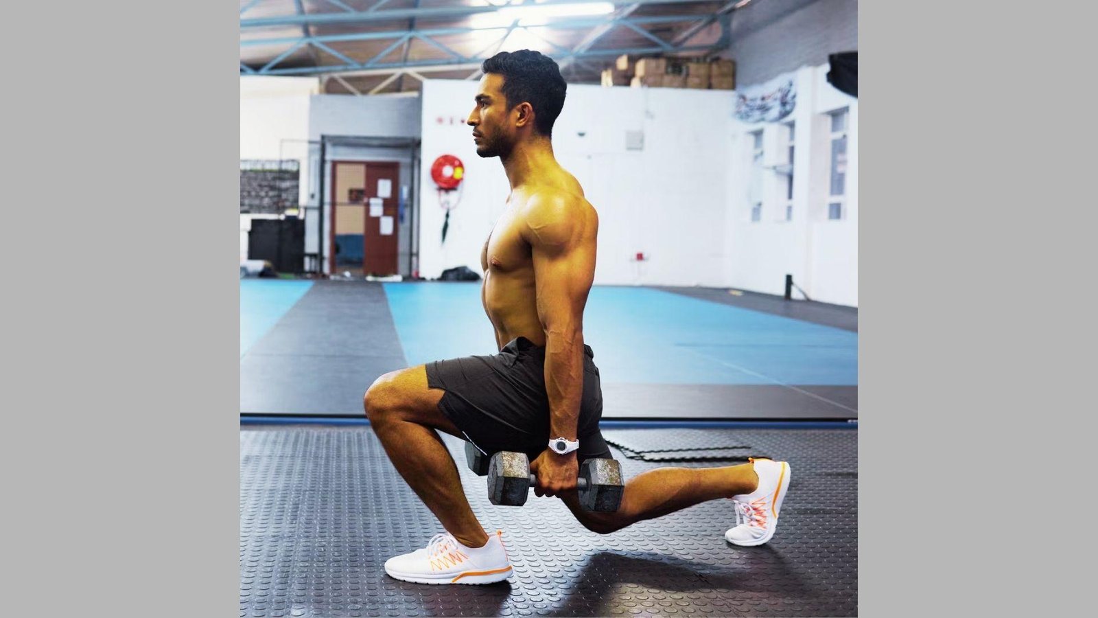 Shirtless muscular man doing dumbbell lunge in gym wearing black shorts, white sneakers holding heavy dumbbells.