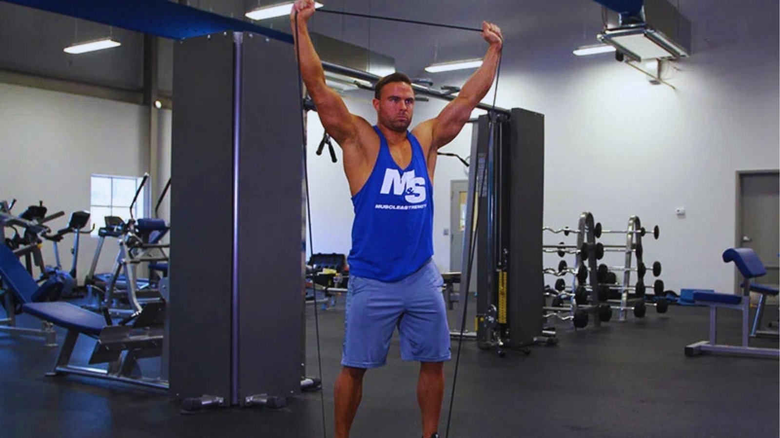 Muscular man doing overhead resistance band pull apart stretch in gym for shoulders and back.