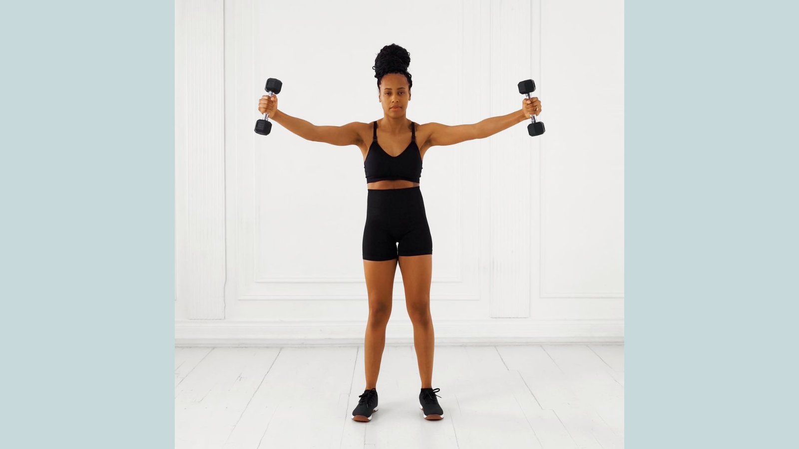 Woman in black sports bra and shorts doing standing dumbbell lateral raises with arms out.