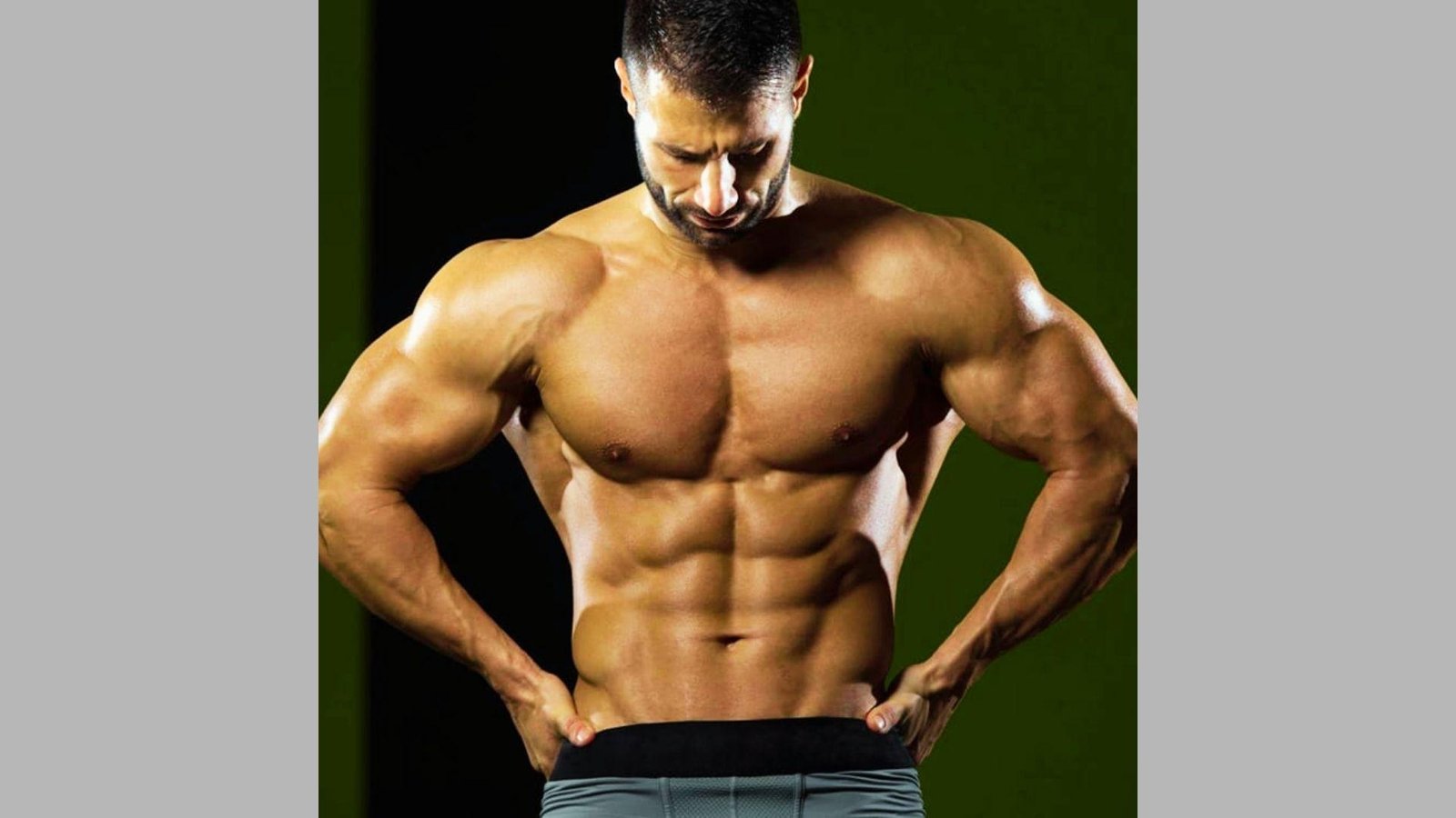 Shirtless muscular man with defined abs and arms posing with hands on waist against dark background.