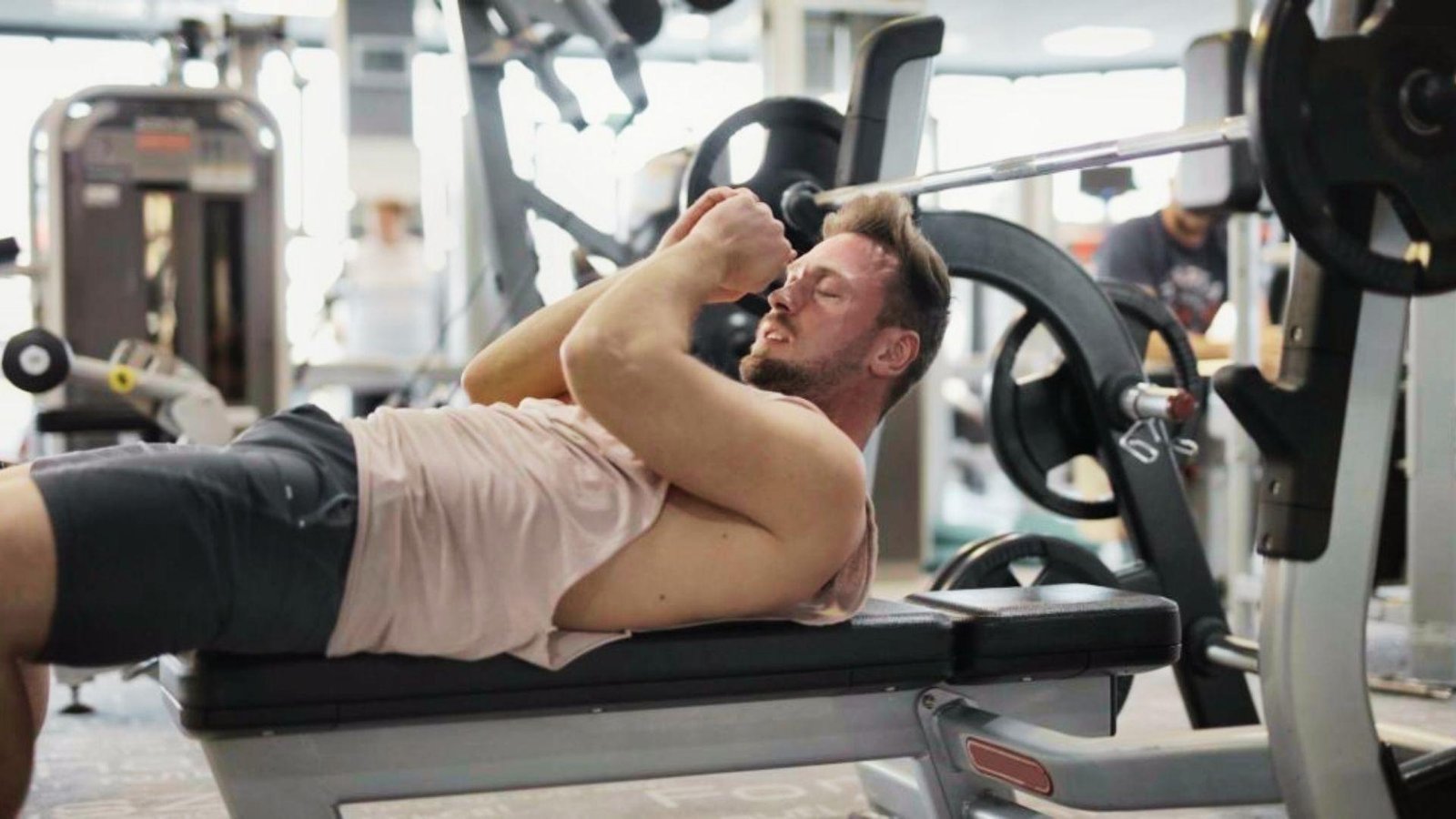 Man in pink tank top lying on bench holding arm in pain near barbell rack in gym.