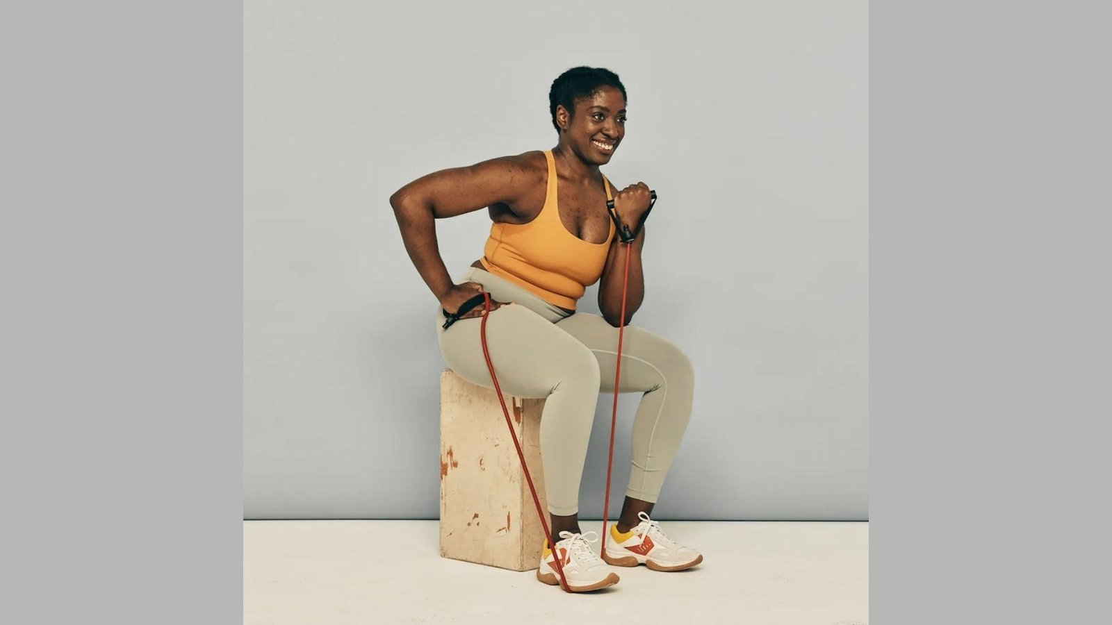 Smiling woman in yellow top sitting on wooden box doing resistance band bicep curl with red band.