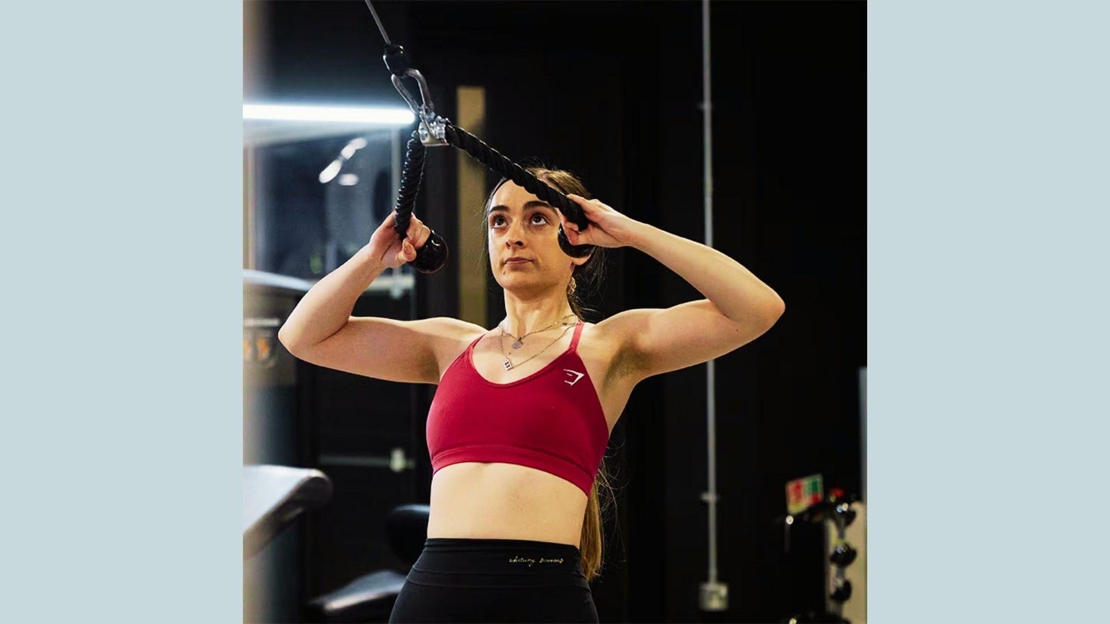 Woman in red sports bra doing cable face pull with rope attachment in gym pulling toward face.