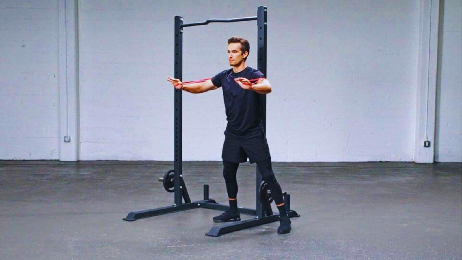 Man doing resistance band chest fly with red mini band in front of squat rack.