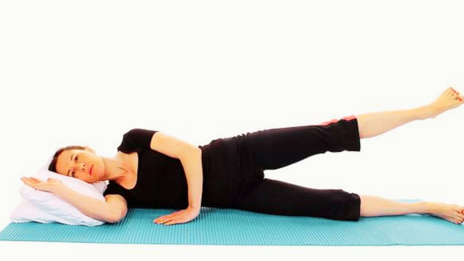 Woman in black outfit lying on side on teal yoga mat doing leg raise with head resting on white pillow.