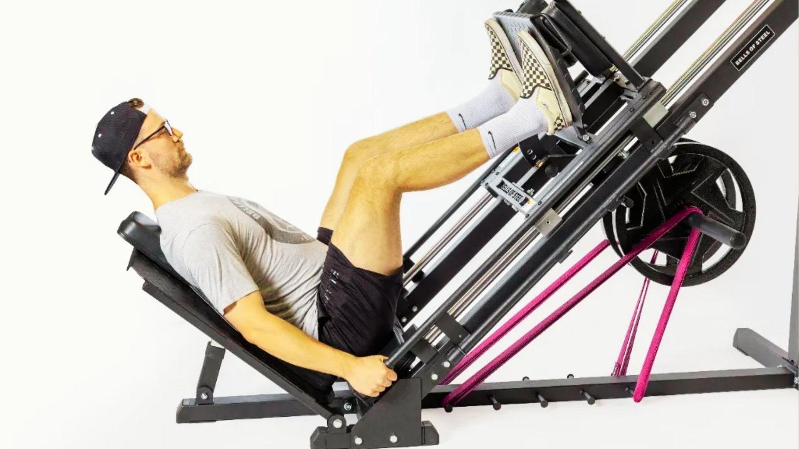 Man in gray shirt, black shorts and cap using leg press machine with pink resistance bands attached.