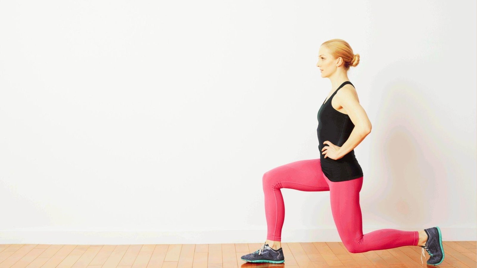 Blonde woman in black tank and pink leggings doing forward lunge on wooden floor against white wall.