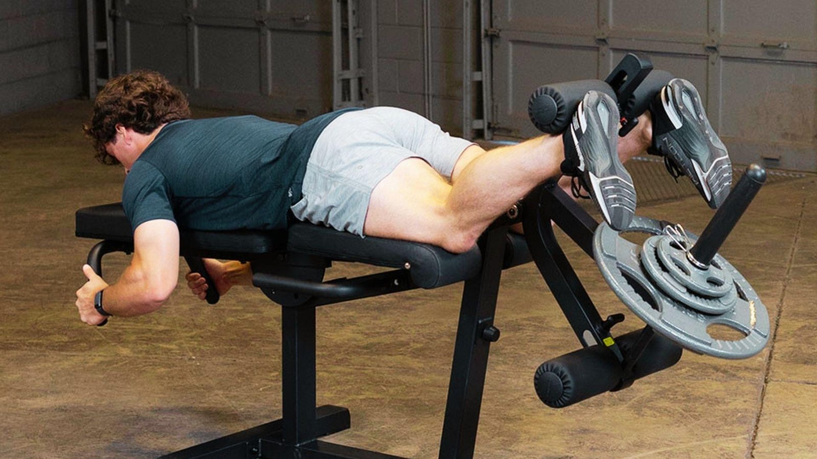 Man in green shirt and gray shorts lying face-down on plate-loaded leg curl machine lifting weights with legs.
