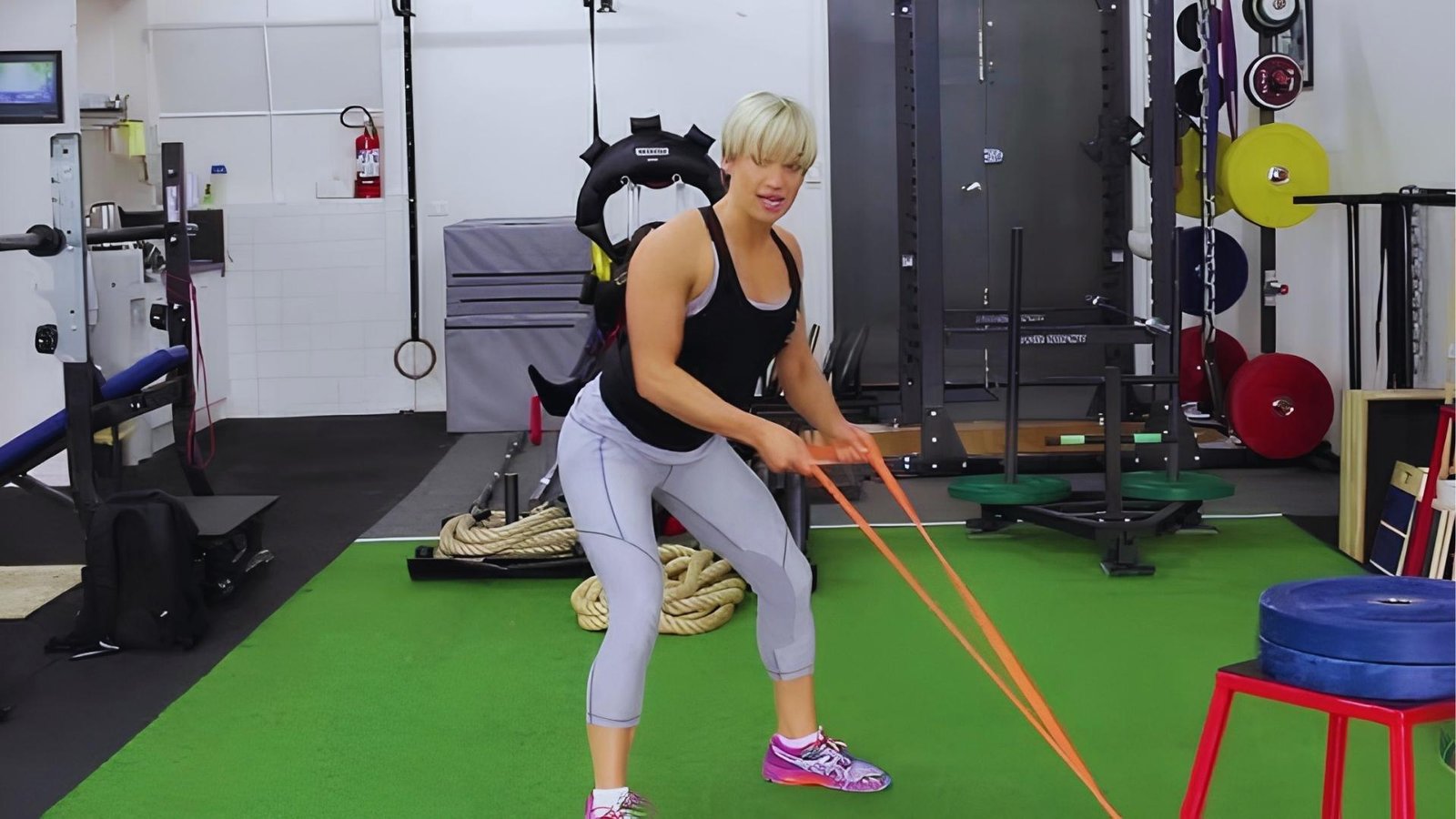 Woman with short blonde hair doing resistance band bent over rows with orange long band in gym.