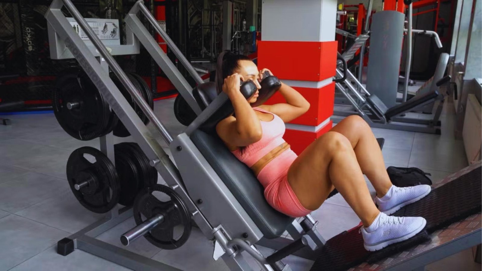 Woman in pink activewear doing hack squat machine exercise for glutes and quads.