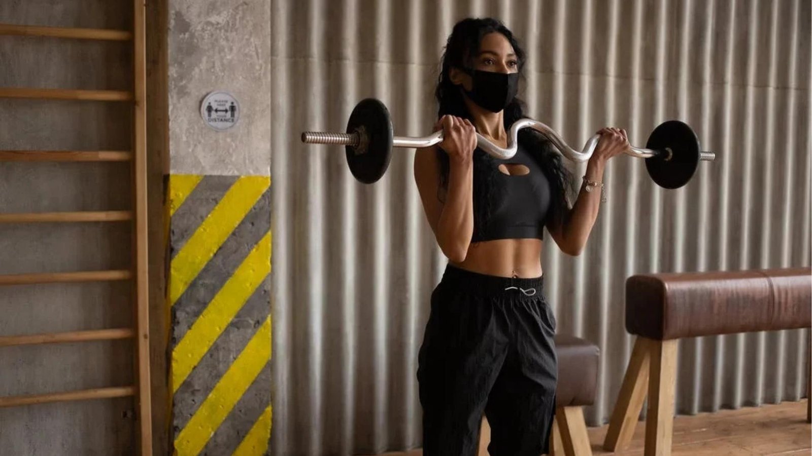 Woman in gym doing EZ bar bicep curls wearing face mask, strength training with barbell.