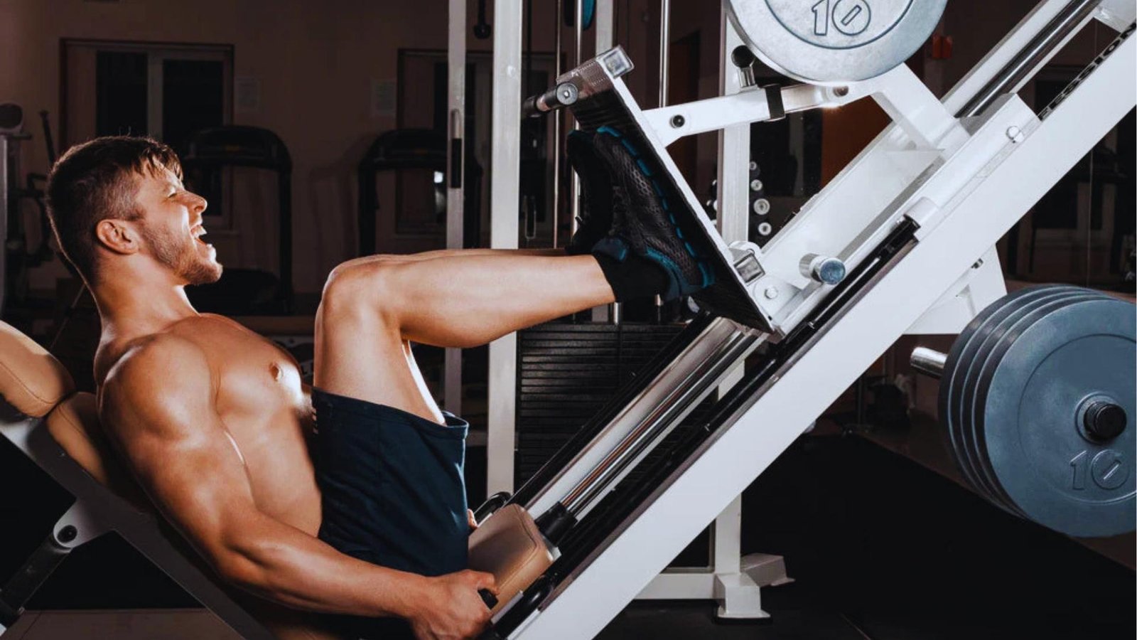 Muscular shirtless man straining on leg press machine with multiple plates, leg day workout.